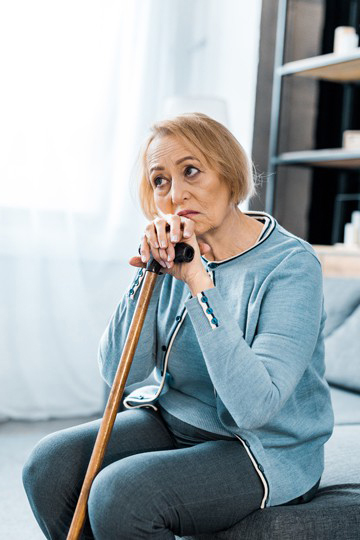 Elderly woman sitting on a couch resting on a wooden cane looking pensive.