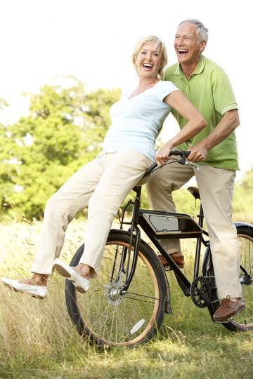 Senior couple laughing and having fun riding a bicycle together in a sunny park.