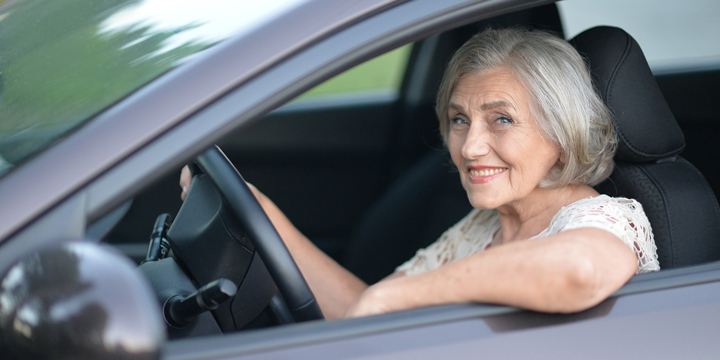 Smiling older woman sitting in a grey car with one hand on the wheel and one arm resting on the open window frame