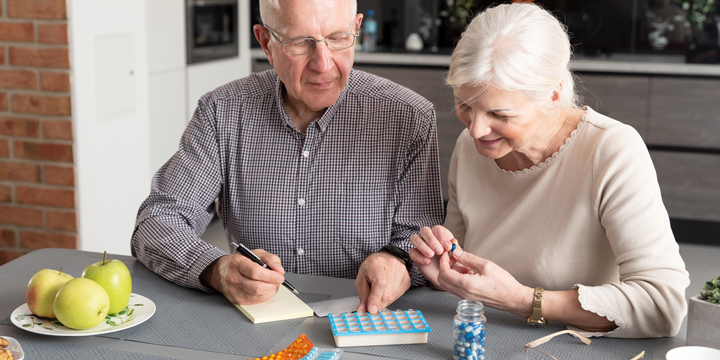 Older man and woman sitting together at a kitchen table sorting pills into a box and making notes on a yellow pad