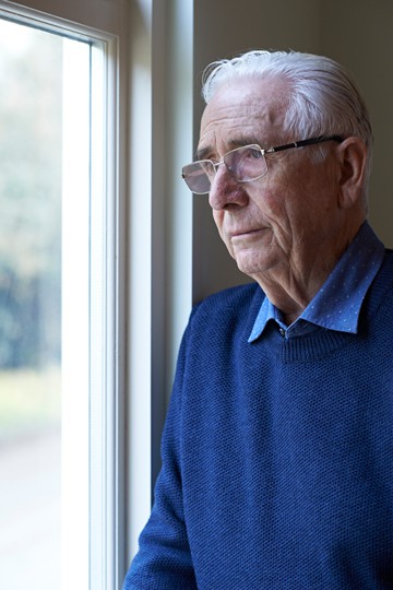 Elderly man with glasses looking pensively out a window while standing indoors