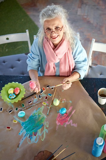 Smiling elderly woman with glasses and a pink scarf painting with acrylics on a canvas at a table with art supplies and a coffee cup.