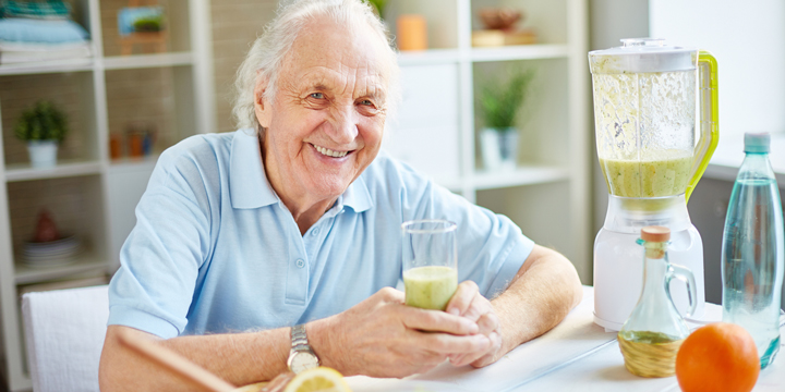 Smiling older man holding a glass of green smoothie while sitting next to the blender that was used to make it