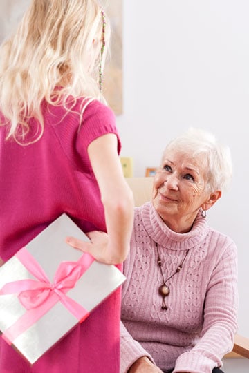 Young girl offering a gift to an elderly woman, who looks at her with a smile in a brightly lit room.