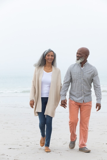 Smiling older couple holding hands, wearing long-sleeved shirts and pants, and walking on a sandy ocean beach