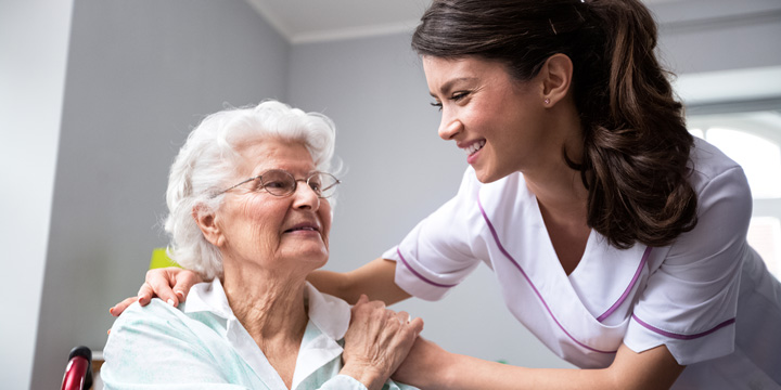 Smiling woman in a medical uniform leaning forward to embrace an older woman who is sitting in a wheelchair