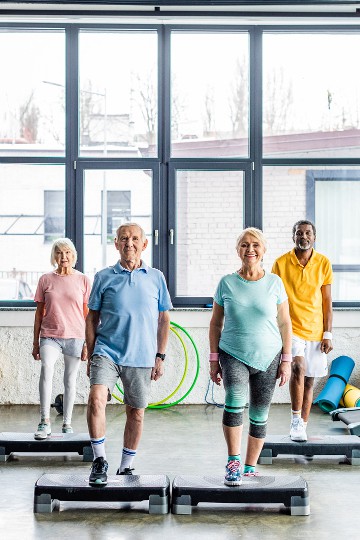Group of senior adults standing on aerobic steppers in a gym class, smiling, with exercise equipment in the background.