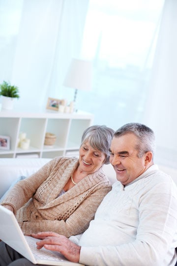 Senior couple smiling and looking at a laptop together in a bright living room setting.