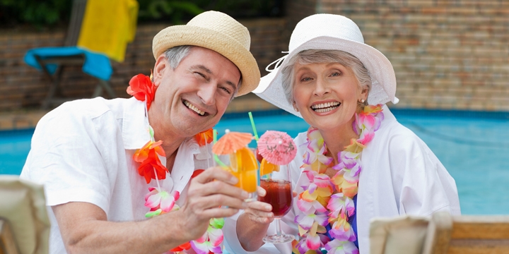 Smiling older couple sitting by a pool wearing hats and flower necklaces while holding drinks with little umbrellas in them