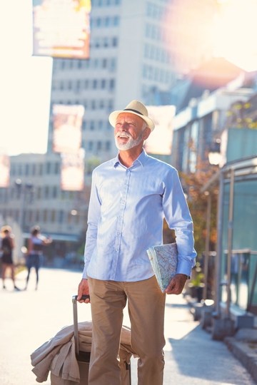 Smiling man with a grey beard and a hat standing on a city street holding a map and pulling a rolling suitcase