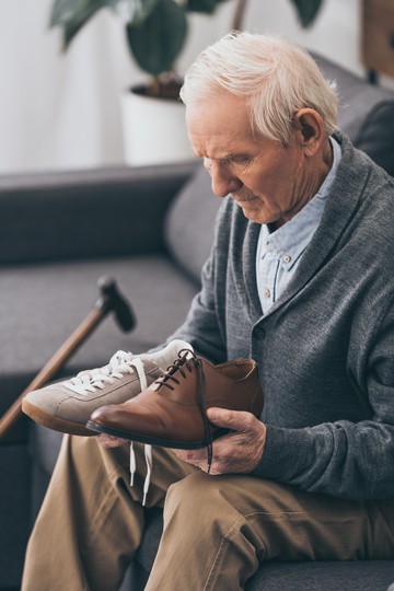 Elderly man sitting in a shoe store trying to choose between two shoes, cane resting nearby.