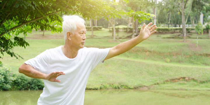Older man in loose white clothing doing tai chi in a park, leaning slightly forward with one arm extended