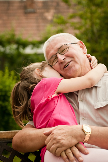 Young girl in a pink shirt hugging a smiling older man with eyeglasses while they sit on a bench outside