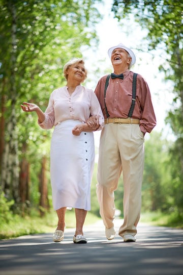 Elderly couple walking and laughing together in a sunny park-lined pathway