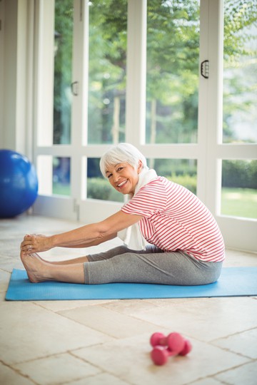 Senior woman smiling while doing stretching exercise on a yoga mat indoors with windows in the background and dumbbells on the floor.