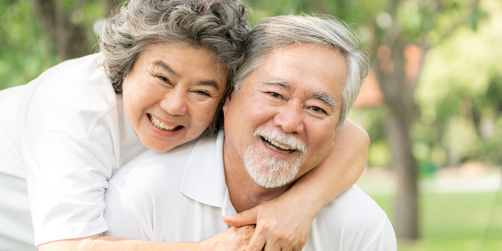 Smiling older woman behind smiling older man embracing him with her head resting on his shoulder