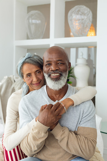 Woman sitting behind smiling man with her arms wrapped tightly around him and their hands clasped together on his chest