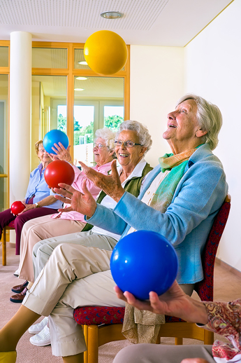 A female senior sitting in a chair joyfully throwing a yellow ball into the air above her