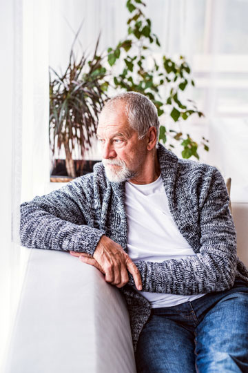 Senior man with beard wearing sweater looking out window while sitting indoors
