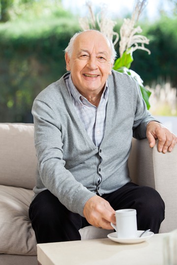 Elderly man smiling sitting on couch holding a cup of coffee with garden view in background