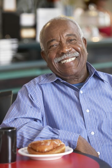 Smiling elderly African American man in blue striped shirt sitting in diner with a donut on a plate.