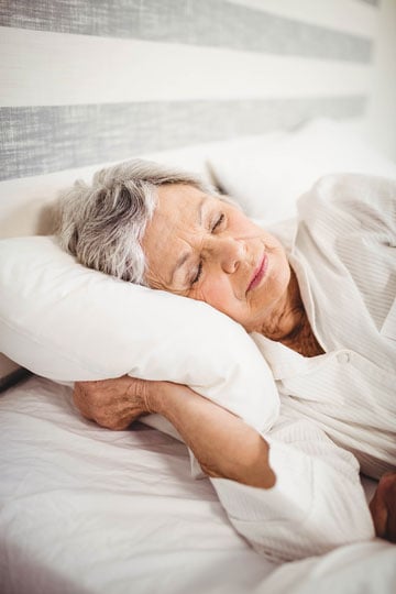 Elderly woman with gray hair sleeping peacefully in a white bed.