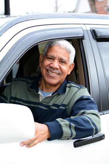 Smiling older man wearing a blue-and-green-striped collared shirt sitting in a car and leaning one arm out the open window