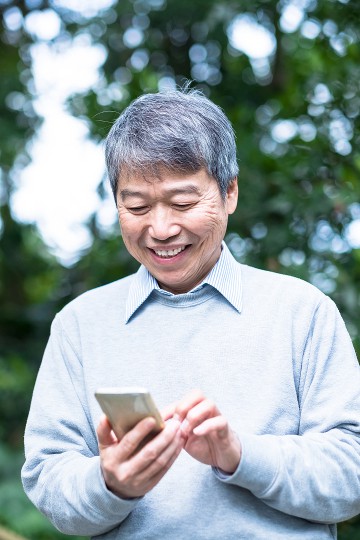 Senior man smiling while using smartphone outdoors with greenery in the background