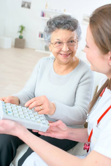 Smiling older woman with grey hair and glasses reaching for a pillbox being handed to her by a young healthcare professional