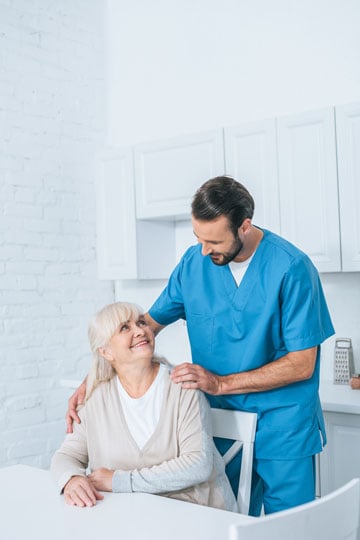 Smiling elderly woman sitting at a table looking up at a male nurse in scrubs who is comforting her with a gentle hand on her shoulder in a bright kitchen.