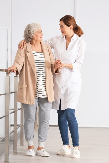 Elderly woman with gray hair being assisted by female healthcare professional in white coat as she walks with handrail support.
