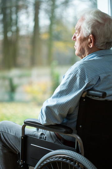 Older man in a collared shirt and gray pants sitting in a wheelchair and looking outside through a floor-to-ceiling window