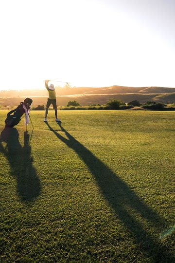 Golfer swinging club on green at sunset with long shadow.
