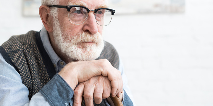 Sitting older man with a worried expression, holding a cane with both hands and resting his chin on his hands