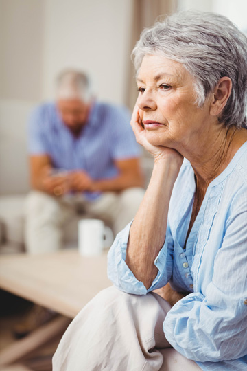 Frowning older woman sitting and resting her chin on one hand, with an older man in the background