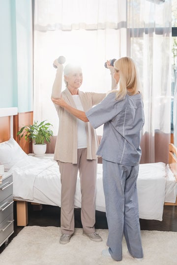 Elderly woman exercising with dumbbells assisted by a female nurse in a sunny room with curtains and a bed in the background