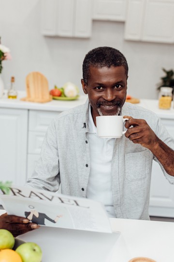 Smiling man enjoying coffee while reading a travel magazine in a kitchen setting.