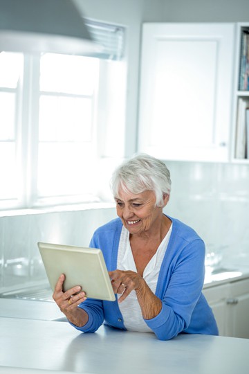 Elderly woman with white hair smiling while using a tablet in a bright room