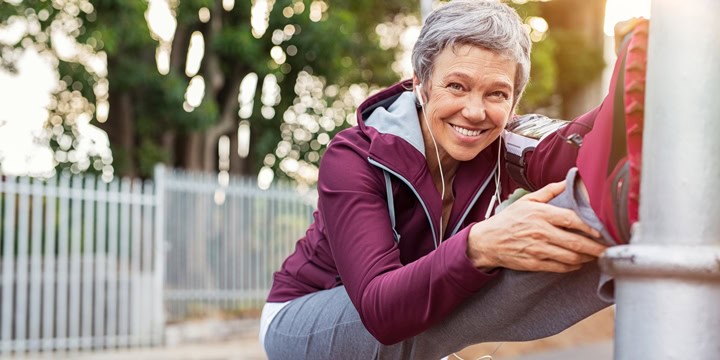 Smiling older woman wearing a tracksuit and headphones and stretching by propping a foot on a post and reaching for it