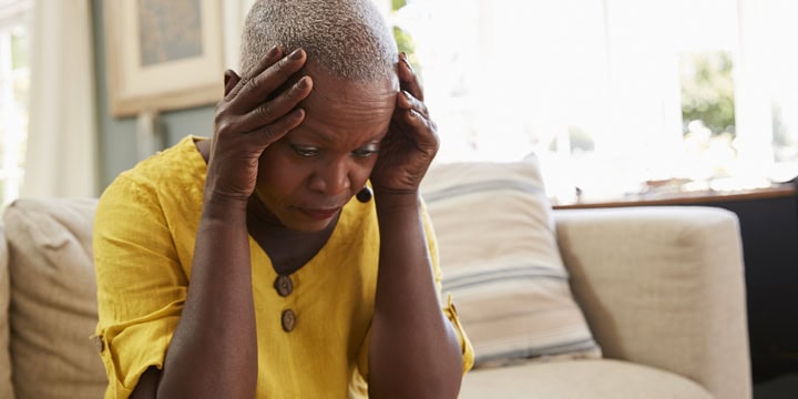 An older woman in a yellow dress sitting on a couch and holding her hands to her temples