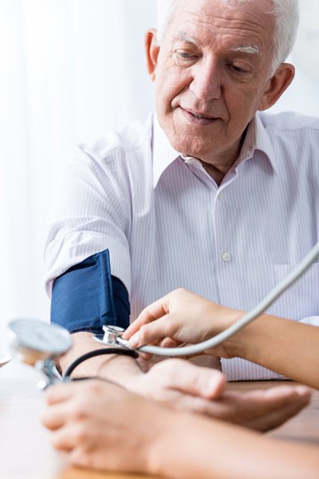 An older man in a white button-up shirt wearing a blood pressure cuff while an unseen person holds a stethoscope to his arm