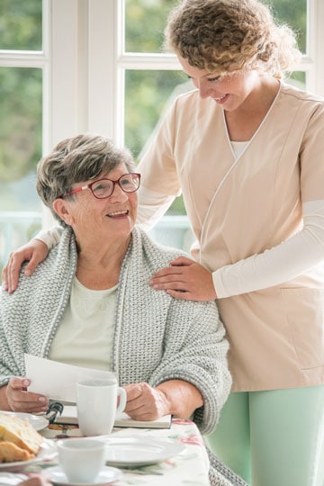 Smiling elderly woman with glasses sitting and smiling with a female nurse in a sunlit room with breakfast on the table.