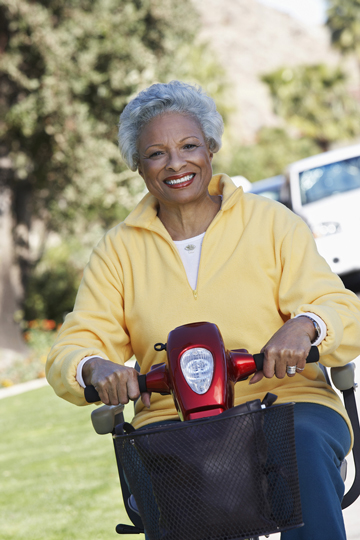 Senior woman with gray hair smiling while riding a red mobility scooter outdoors