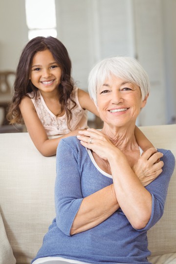 Smiling elderly woman with granddaughter hugging from behind on couch at home