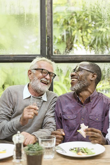 Two senior men laughing and enjoying a meal together indoors by a window