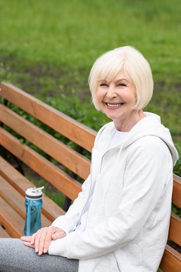 Smiling older woman with a water bottle and a white hoodie sitting on a park bench