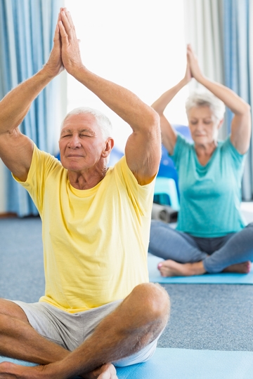 Gray-haired woman and man sitting cross-legged on yoga mats with their eyes closed and their hands together above their heads