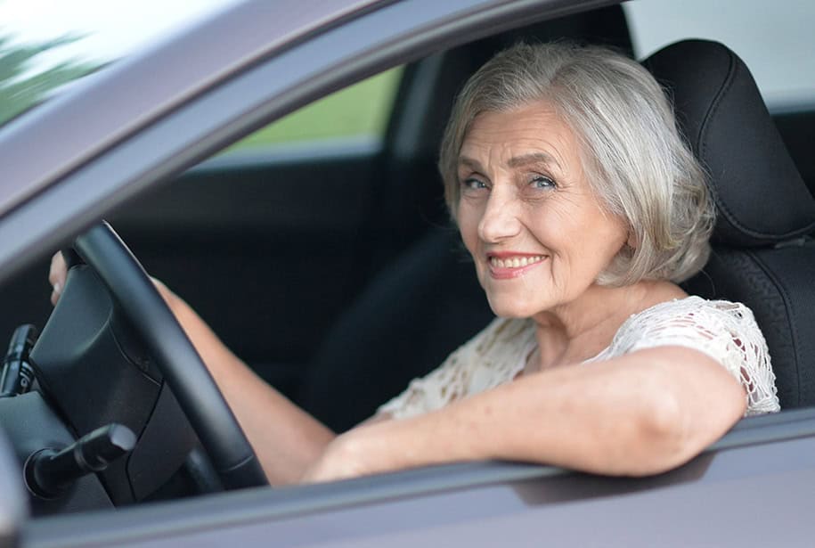 Smiling older woman sitting in a grey car with one hand on the wheel and one arm resting on the open window frame