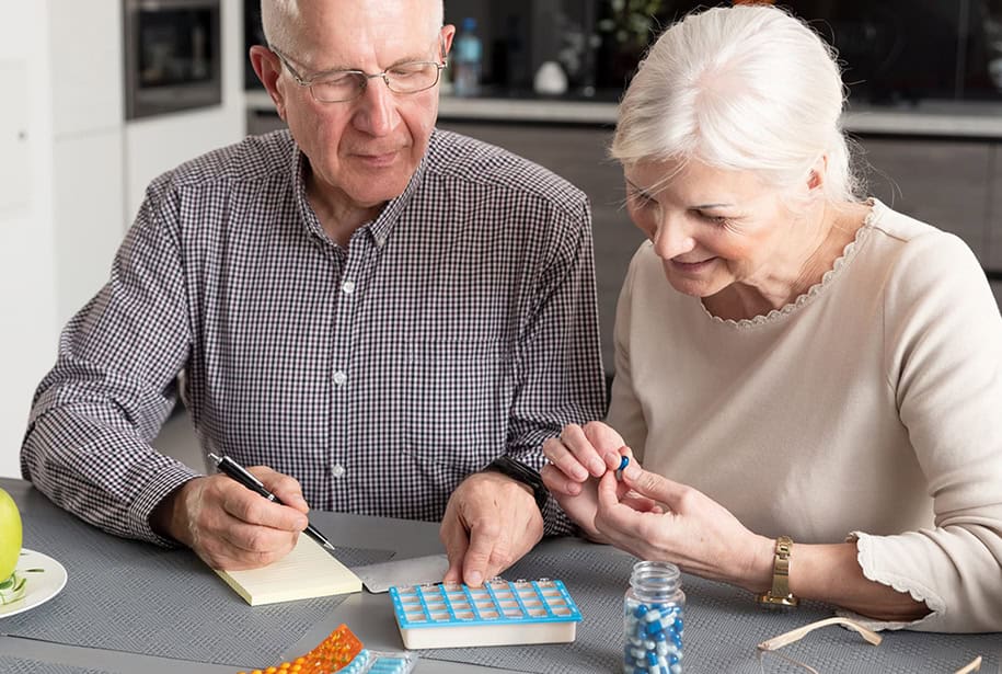 Older man and woman sitting together at a kitchen table sorting pills into a box and making notes on a yellow pad