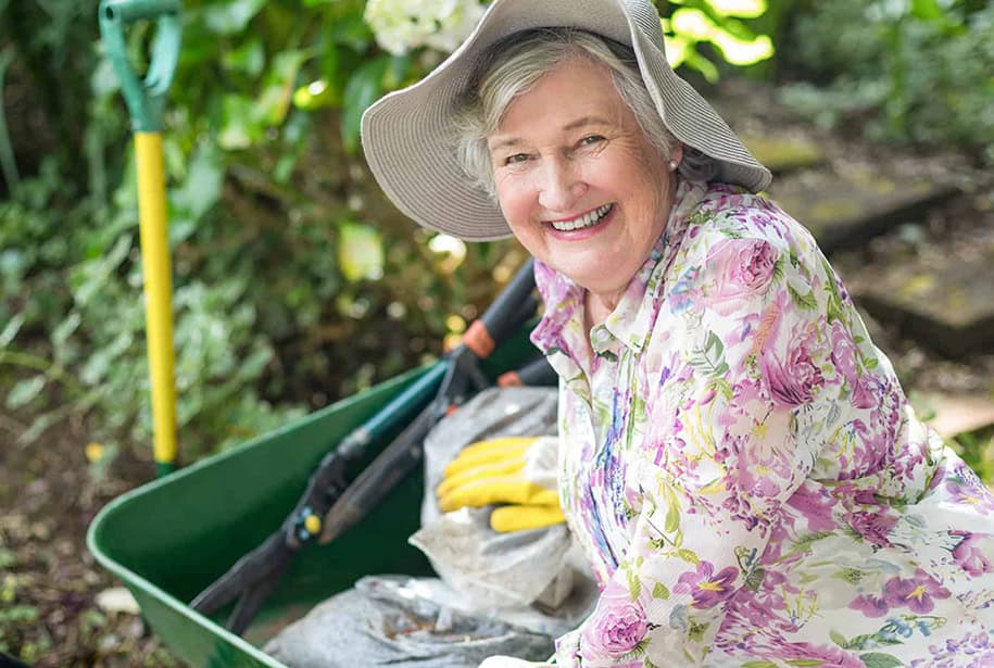 Smiling senior woman wearing a sun hat and floral shirt, gardening with a wheelbarrow and tools outdoors.